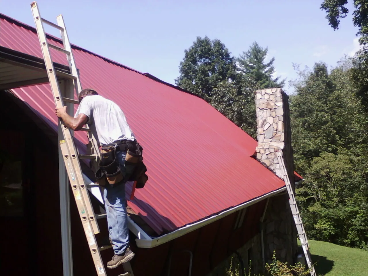 Roofer on a ladder beside a red metal roof and stone chimney — Soffit & Fascia Repair by Roofing Brothers in La Plume